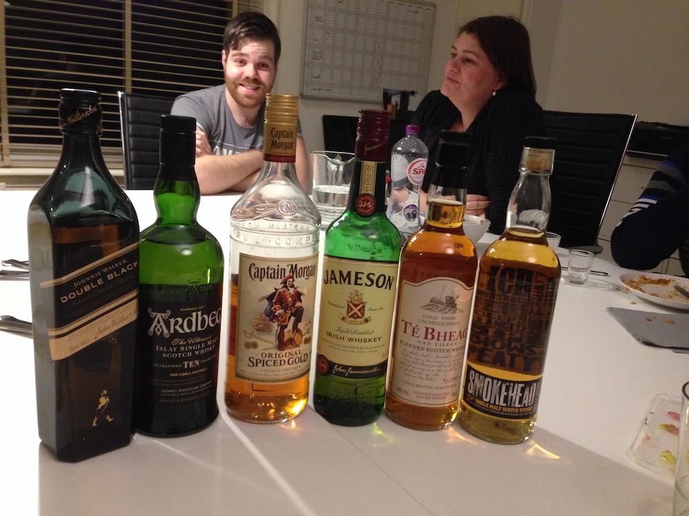 Five bottles of whisky and one bottle of rum lined up at an #a11y meeting, Tim and Iacobien in the background.